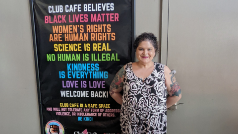 Bela stands with a large poster at Club Cafe in Boston, Massachusetts. The poster text reads: Club Cafe believes Black lives matter, women’s rights are human rights, science is real, no human is illegal, kindness is everything, love is love, welcome back! Club Cafe is a safe space and will not tolerate any form of aggression, violence, or intolerance of others. Be kind!