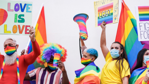 A group of diverse people are wearing brightly colored clothes featuring rainbow colors.  They’re responsibly wearing face masks while carrying signs, megaphones, and rainbow cups. The signs say: Love is Love, LGBT, and Fight for Equality.