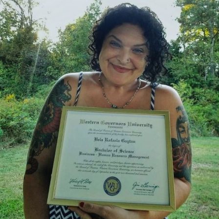 Bela stands outside, holding her Bachelor's Degree diploma.