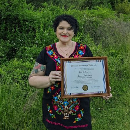Bela stands outside, wearing a black, floral, Mexican dress, holding her Master's Degree diploma.
