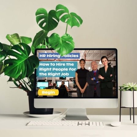 An all-in-one computer sits on a white desk with plants on each side. On the computer screen is “HR Hiring Policies, how to hire the right people for the right job” with a begin button below. To the right are three smiling people dressed professionally with an office environment in the background.